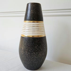May include: A tall, decorative vase with a speckled black base and a cream-colored, horizontally striped midsection. Gold bands accent the top and middle sections. The vase has a tapered shape and a dark speckled top.