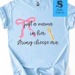 Könnte beinhalten: Hellblaues T-Shirt mit dem Text "just a mama in her string cheese era". Ein rosa Schleife und eine Grafik von Käsefäden sind ebenfalls auf dem Shirt abgebildet. Das Shirt hat kurze Ärmel und einen Rundhalsausschnitt.