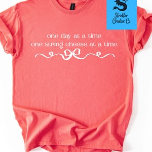 May include: Coral-colored t-shirt with white text: "one day at a time, one string cheese at a time." A decorative ribbon design is below the text. The upper right corner features a blue square with a black dragon logo and "Strickler Creative Co." text.