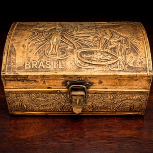 May include: A vintage brass-colored box with a hinged lid and clasp. The box is adorned with embossed designs, including figures and the word "BRASIL". The box has a rectangular shape with a curved top and is likely used for storage.
