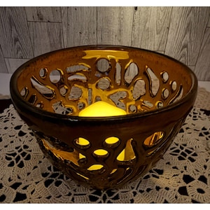 May include: A decorative amber-colored glass bowl with an intricate, openwork design. The bowl has a round shape and contains a yellow candle, creating a warm glow. The bowl sits on a white, lacy doily, adding a touch of elegance.