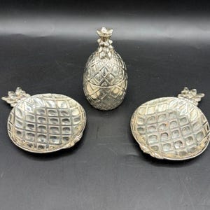 May include: A set of three silver-toned metal items consisting of one pineapple-shaped lidded container and two matching circular dishes, each featuring a textured, grid-like pattern resembling the exterior of a pineapple.