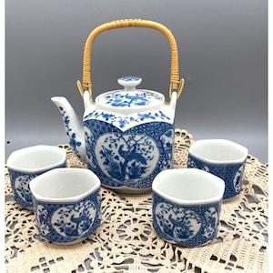 May include: A white and blue ceramic tea set with a teapot and four cups. The teapot has a woven handle and floral and bird designs. The cups have a similar design. The set is on a lace doily.