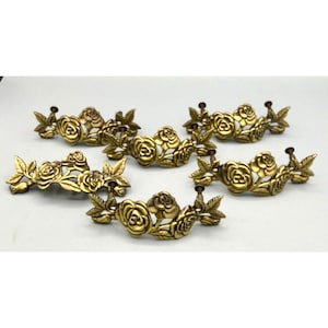 May include: Six antique brass drawer pulls with a rose and leaf design. The pulls are arranged in a semi-circle, each featuring detailed floral elements. The brass has a warm, aged patina, suggesting vintage craftsmanship. These decorative pulls would be suitable for a dresser or cabinet.