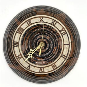 May include: Round ceramic wall clock with a spiral design in shades of brown, black, and cream. The clock face features Roman numerals and gold-colored hands. The clock has a textured, ridged border.