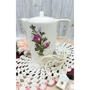 May include: White ceramic electric teapot with a lid, featuring a floral design of pink roses and green leaves. The teapot has a handle and a spout, and a white electrical cord is coiled nearby. A vintage kitchenware item.