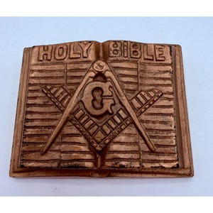 Puede incluir: Una placa de metal color cobre con forma de libro abierto con las palabras HOLY BIBLE en relieve en la parte superior. Centrado en las páginas hay un símbolo masónico en relieve de una escuadra y un compás con la letra G en el medio.