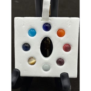 May include: A white square pendant with a central oval opening and eight colorful, round gemstones. The gemstones are blue, orange, red, purple, yellow, and green. A white ribbon is attached at the top.