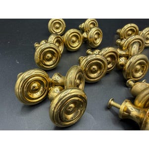 May include: A collection of circular brass cabinet knobs featuring a stepped, concentric ring design. Each knob has a polished metallic finish and a central raised dome, with a threaded metal screw extending from the back for installation.