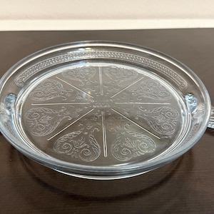 May include: Clear glass serving tray with a decorative etched floral design on the interior. The tray has a circular shape with a raised rim and two small handles on either side. The tray is sitting on a dark brown surface.