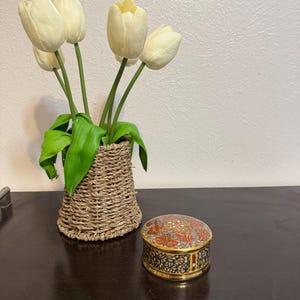 May include: A woven basket containing five cream-colored artificial tulips with green leaves, positioned next to a small, round, lidded metal box featuring a detailed red, gold, and black geometric pattern on the lid and a black and white floral design.
