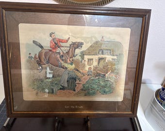 Op de afbeelding: Ingelijste vintage prent die een vossenjachtscène uitbeeldt. Een man te paard springt over een vrouw in een tuin. Een huisje en honden staan op de achtergrond. De tekst "Got the Brush" staat onderaan.