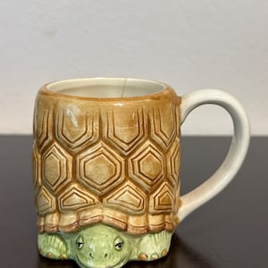 Może przedstawiać: Ceramiczny kubek z motywem żółwia. Kubek ma brązowy, teksturowany wzór skorupy żółwia po bokach i zieloną twarz żółwia u podstawy. Uchwyt jest biały, a wnętrze kremowe.