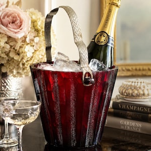 Könnte beinhalten: Ein roter Champagner-Eimer aus Glas mit einem silbernen Griff, gefüllt mit Eis und einer Champagnerflasche. Der Eimer hat ein geripptes Design. Zwei Champagnergläser befinden sich im Vordergrund und ein Blumenstrauß im Hintergrund.