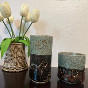 May include: Two ceramic cups with a crackle glaze in teal and brown tones. The taller cup has a decorative design, and the shorter cup has cut-out shapes. A woven basket holds white tulips.