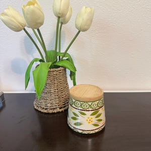 May include: A decorative ceramic jar with a wooden lid, featuring a green and yellow floral pattern. Beside it, a woven basket holds a bouquet of cream-colored tulips. The arrangement is set on a dark wooden surface.