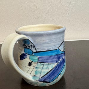 May include: Hand-painted ceramic mug with a light blue rim and handle. The mug features a design of blue and turquoise shapes with the number "N477" on the side. The interior is a cream color.