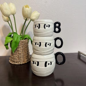 Peut inclure: Trois tasses en céramique blanche empilées, décorées de visages de momies avec des yeux noirs et des motifs de bandages. Les anses sont noires et les tasses forment le mot "BOO".