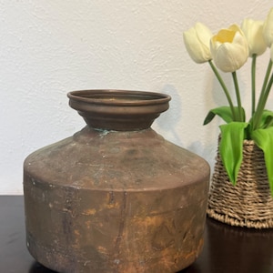 May include: A vintage copper water pot with a wide, rounded body and a flared rim. The pot has a weathered, aged appearance with visible patina and discoloration. The pot is displayed on a dark surface, with a wicker basket and white tulips in the background.