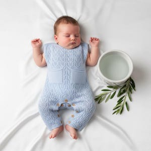 May include: A sleeping baby wearing a light blue knitted romper with wooden buttons. The romper has a textured pattern and two front pockets. A white bowl with water and green leaves are next to the baby on a white surface.