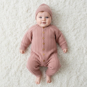 May include: A baby wearing a dusty rose-colored knitted romper and matching beanie. The romper features a ribbed design and wooden buttons down the front. The baby is lying on a white, fluffy rug.