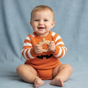 May include: A baby wearing an orange and white striped pumpkin-themed sweater and holding a wooden pumpkin-shaped teether. The sweater has a black jack-o'-lantern face design on the front. The baby is sitting on a blue surface.