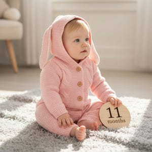 May include: A baby wearing a pink knitted bunny-eared hooded jacket and matching pants, sitting on a gray rug. The baby holds a wooden disc with the number "11" and the word "months" printed on it. The outfit has wooden buttons.