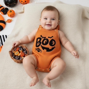 Può includere: Un body arancione per neonati con una faccia di zucca nera e la scritta "BOO" stampata sul davanti. Il bambino è sdraiato su una coperta color crema, con un piccolo cestino di caramelle nelle vicinanze. Abbigliamento per bambini a tema Halloween.