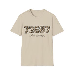 Puede incluir: Camiseta beige con el número "72687" en estampado de leopardo, contorneado en negro. Debajo de los números, el texto "Yellville Arkansas" está escrito en cursiva. La camiseta tiene mangas cortas y cuello redondo.
