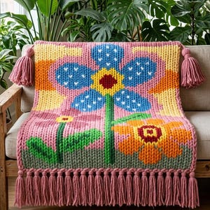 May include: A chunky knit throw blanket featuring a large blue flower with a yellow center, a smaller pink flower, and an orange flower on a pink background. The blanket has thick pink tassels on the top corners and a long pink fringe along the bottom.