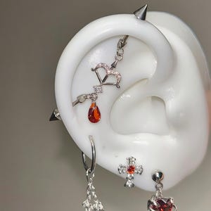 Pode incluir: Uma variedade de brincos prateados, incluindo uma corrente pendurada, um aro, uma cruz com uma gema vermelha e um brinco em forma de coração com uma gema vermelha. Um brinco de arco e flecha e um brinco em forma de gota com uma gema vermelha também são visíveis.