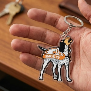 May include: A clear acrylic keychain featuring a cartoon dog with the word "SMOKEY" in orange. The dog has black, white, and orange markings and is attached to a silver chain and keyring. The keychain is held in a hand.
