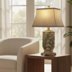 May include: A decorative table lamp with a beige lampshade and a detailed base featuring a leaf design. The lamp sits on a wooden side table next to a cream-colored armchair. The room has a neutral color palette.