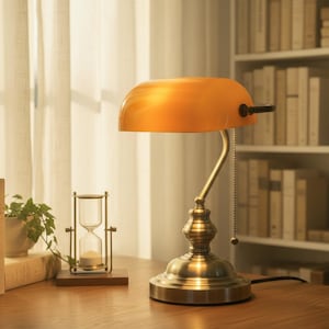 May include: A banker's lamp with a curved amber glass shade and a metallic brass-toned base. The lamp features a pull chain switch on the side, a curved neck, and a circular pedestal base, emitting a warm glow across the surface.