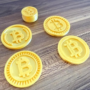 Pode incluir: Várias moedas Bitcoin amarelas, algumas empilhadas, com o símbolo Bitcoin em relevo. As moedas estão dispostas sobre uma superfície de madeira, destacando o seu design e cor. Cada moeda tem cerca de 4 cm de diâmetro.