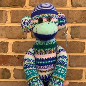 May include: A stuffed monkey toy crafted from knit fabric featuring a colorful fair isle pattern in shades of blue, green, pink, and white. The monkey has two black button eyes, a light green muzzle, and a small white tag attached to its ear.