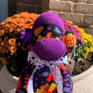 May include: A stuffed monkey toy crafted from fabric with a vibrant floral pattern in purple, orange, yellow, and black. The monkey features two dark circular button eyes and a white ribbon tied around its neck, decorated with colorful polka dots.