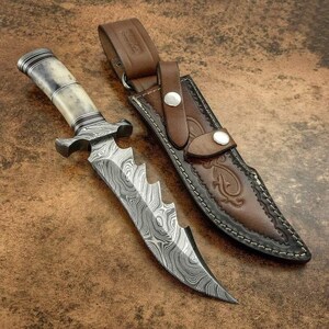 Può includere: Un coltello a lama fissa con lama in acciaio Damasco dotata di dorso seghettato e manico chiaro con venature scure. Il coltello è accompagnato da un fodero in pelle marrone con cuciture decorative, due bottoni a pressione e un passante.