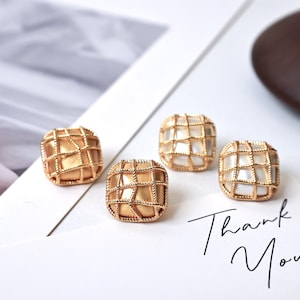 Könnte beinhalten: Vier quadratische, goldfarbene Ohrringe mit einem gitterartigen Design. Zwei Ohrringe haben ein einfarbiges Gold-Aussehen, während die anderen beiden ein weißes, gitterförmiges Innendesign haben. Die Worte "Thank You" sind in Schreibschrift geschrieben.