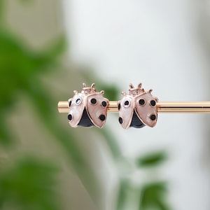 Könnte beinhalten: Ein Paar Ohrstecker in Form von Marienkäfern. Die Ohrringe haben eine roségoldene Oberfläche mit weißen und schwarzen Emaille-Details. Jeder Marienkäfer hat schwarze Punkte und eine schwarze Unterseite. Die Ohrringe werden auf einem goldenen Stab präsentiert.