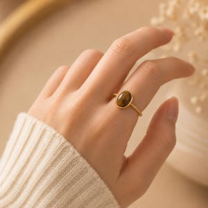 Könnte beinhalten: Ein goldener Ring mit einem ovalen Tigeraugenstein. Der Ring hat ein strukturiertes Band und ist in einer goldenen Lünette gefasst. Der Ring befindet sich an einem Finger, der Hintergrund ist unscharf.