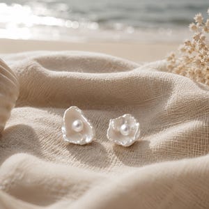 Könnte beinhalten: Ein Paar weiße Ohrringe mit Perlen in der Mitte, geformt wie offene Muscheln. Die Ohrringe liegen auf einem beigen Stoff, im Hintergrund eine Muschel und Trockenblumen. Ein zartes, elegantes Accessoire.