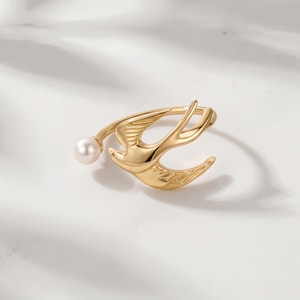 Könnte beinhalten: Ein goldfarbener Ring mit einem stilisierten Vogel im Flug. Der Ring hat ein filigranes Design mit einem kleinen, runden Perlenakzent. Die Flügel des Vogels sind ausgebreitet, und der Ring ist vor einem weißen Hintergrund platziert.