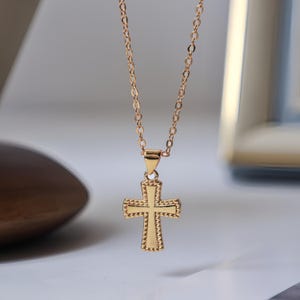 Op de afbeelding: Een goudkleurige ketting met een kruisvormige hanger. Het kruis heeft een gedetailleerd ontwerp met een kralenrand. De ketting is delicaat en ook goudkleurig. De ketting wordt gepresenteerd tegen een zachte, neutrale achtergrond.