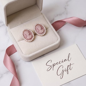 Könnte beinhalten: Ovale Cameo-Ohrringe mit rosa Profil, umgeben von kleinen Perlen, präsentiert in einer Samt-Schmuckschatulle. Ein rosa Band und eine Karte mit der Aufschrift "Special Gift" vervollständigen die Präsentation. Die Ohrringe haben einen goldfarbenen Rahmen.