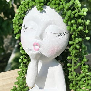 Könnte beinhalten: Weißer Keramik-Blumentopf in Form eines Gesichts mit geschlossenen Augen und einem Kussmund. Das Gesicht hat rosa Wangen und Lippen. Grüne, kaskadierende Kunst-Sukkulenten bilden das Haar. Ein dekoratives Objekt.