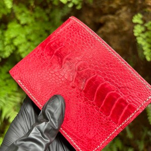 May include: A vibrant red leather wallet with a textured surface and white stitching. The wallet is rectangular and appears to be made of high-quality material. The background is a blurred view of green foliage.