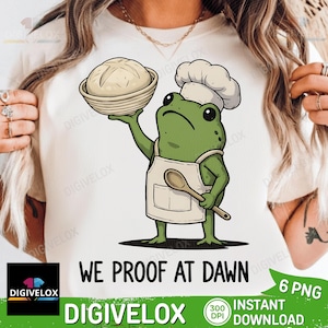 May include: A white t-shirt featuring a graphic of a green frog wearing a white chef hat and apron. The frog holds a round loaf of bread in a basket and a wooden spoon. Below the graphic, the text reads: WE PROOF AT DAWN in black capital letters.
