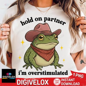 May include: A graphic design featuring a cartoon frog wearing a cowboy hat and a red bandana. The frog is green with yellow accents and is surrounded by yellow starbursts. Text reads "hold on partner" and "i'm overstimulated".