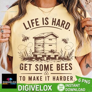 Könnte beinhalten: Ein hellgelbes T-Shirt mit einer dunkelbraunen Grafik eines Bienenstocks, fliegenden Bienen und dem Text "LIFE IS HARD. GET SOME BEES TO MAKE IT HARDER." Das Design enthält kleine florale Elemente an der Basis des Stocks und ein Wabensymbol.